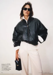 Model wearing dark sunglasses, a black leather bomber jacket, and light trousers holding a small black clutch against a plain white background