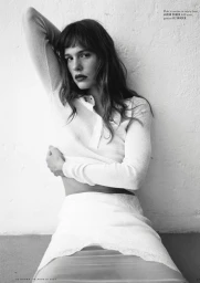Model in a white ribbed polo top and white skirt leaning against a textured wall in a black and white editorial portrait