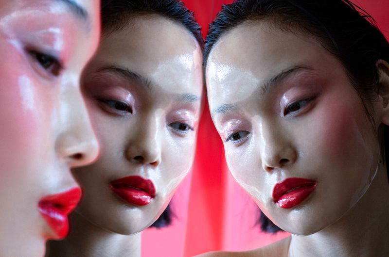 Close-up studio shot featuring an Asian female model with glossy skin, red lipstick, and pink eye shadow, reflected in a mirror.
