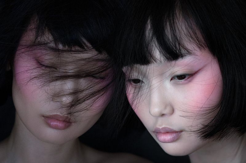 Close-up portrait of two Asian adult females with pink blush makeup and dark hair against a dark background, fashion and beauty.