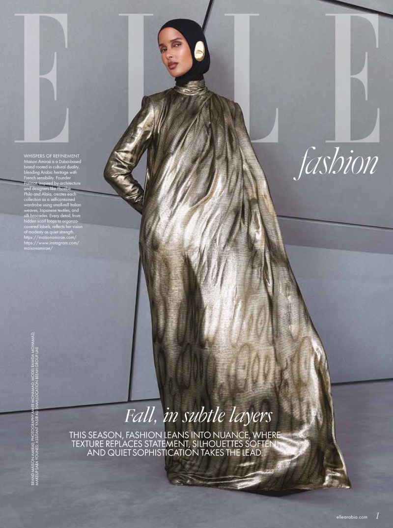 A woman in a gold dress and black headscarf poses for the cover of Elle fashion magazine against a minimalist grey background indoors.