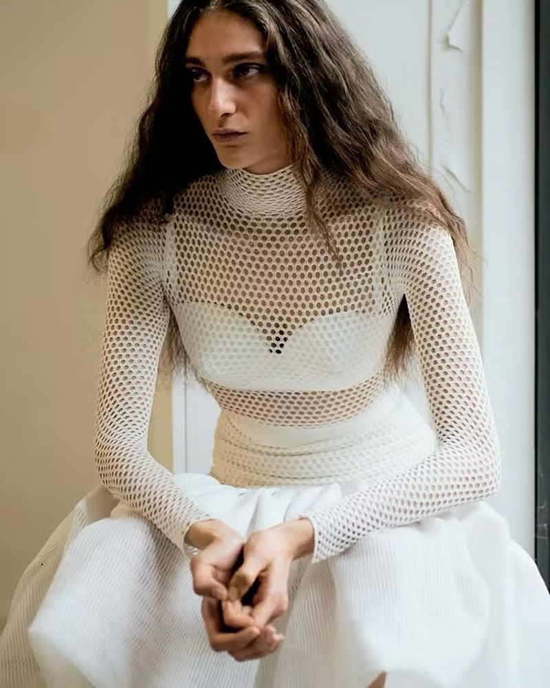 A woman with long brown hair wears a white mesh top and skirt, looking to the side in a studio with natural light and neutral colors.