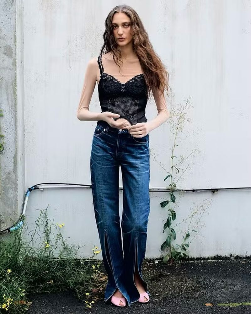 A stylish adult woman in a lace top and blue jeans poses against a textured wall with greenery; fashion, full shot, outdoor daylight.