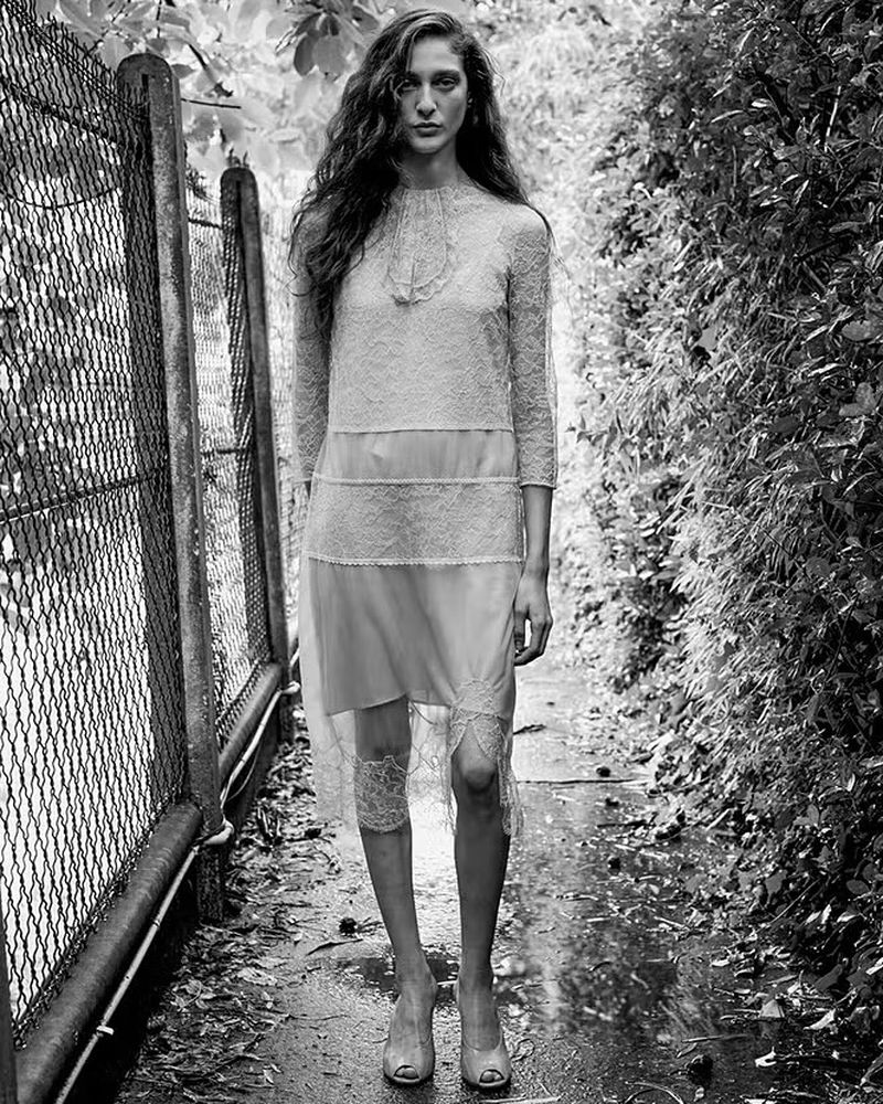 A fashion photo in black and white featuring a slender woman with long hair, wearing a lace dress and high heels, standing on a wet path.