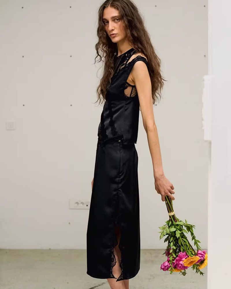 Young woman in a black lace-trimmed satin top and skirt holding a bouquet of flowers against a neutral background, fashion shot.