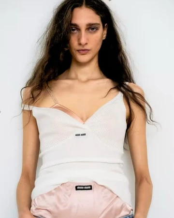 A woman with long brown hair poses indoors, wearing a white knit top with a visible brand logo against a neutral backdrop.