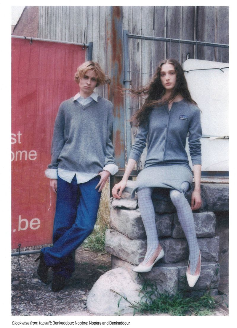 Two fashion models pose outdoors in grey sweaters and blue bottoms. The woman sits on stone steps, the man leans against a red wall.