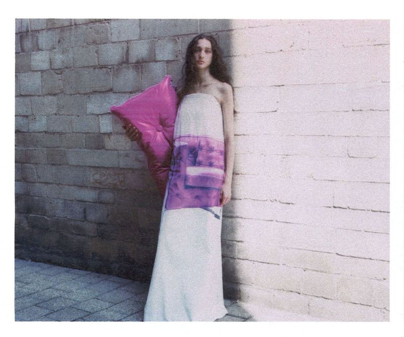 A fashion photo features a young adult woman posing outdoors in a white dress holding a pink pillow against a brick wall backdrop.
