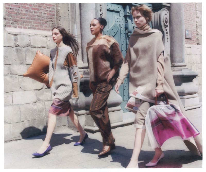 Three fashion models walk along a stone city street in contemporary knitwear, carrying unique bags and purses, photographed in bright daylight.
