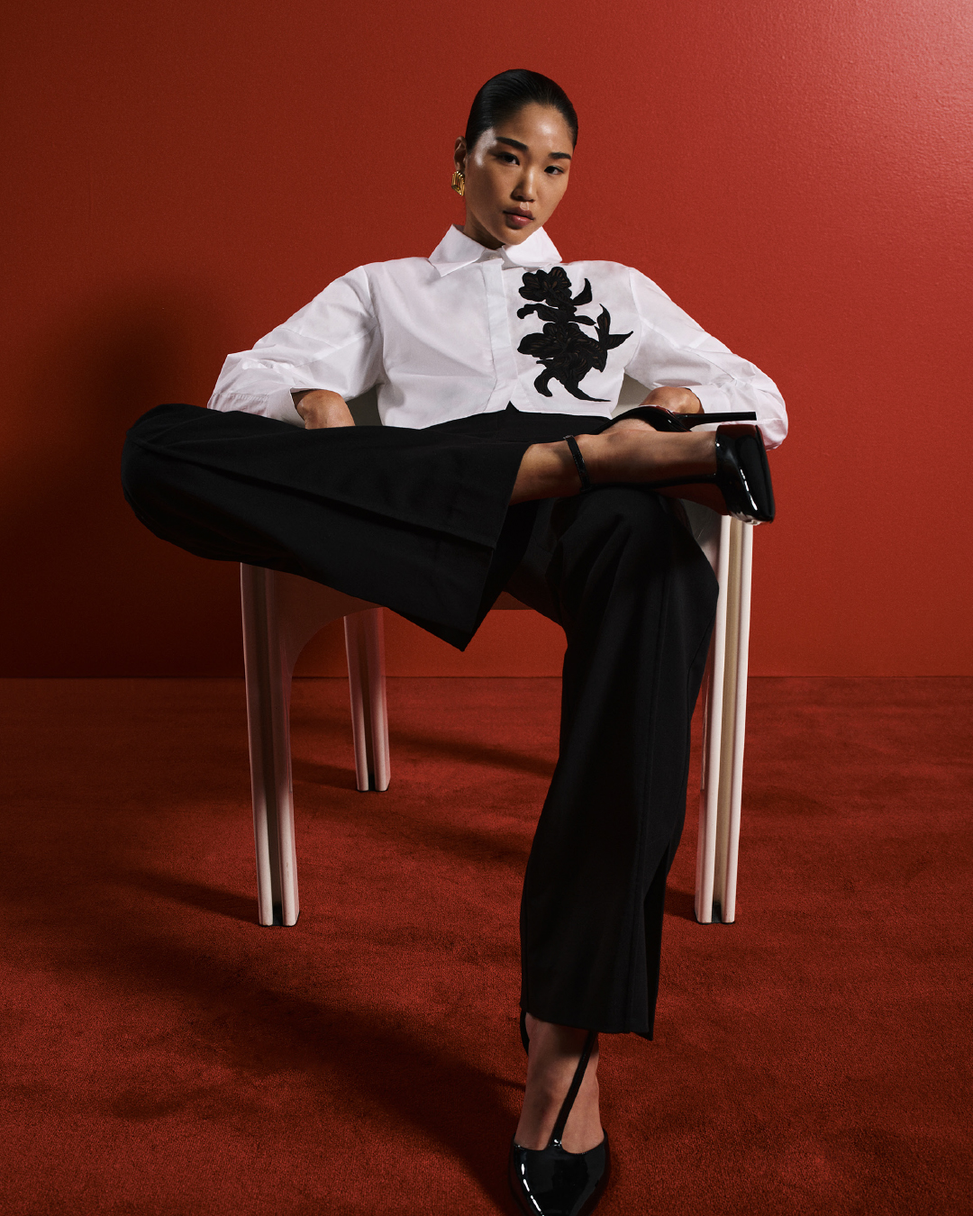Model wearing a white shirt with black applique and black trousers sitting in a chair against a red backdrop
