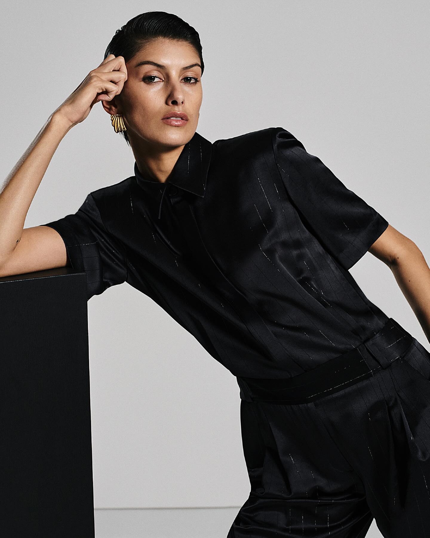 Woman in black satin button-down shirt and pants with gold earrings, posing against a gray background with a black geometric block.