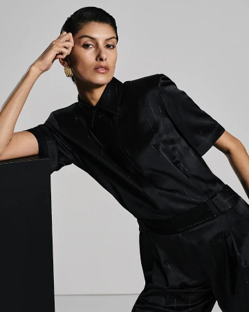 Woman in black satin button-down shirt and pants with gold earrings, posing against a gray background with a black geometric block.