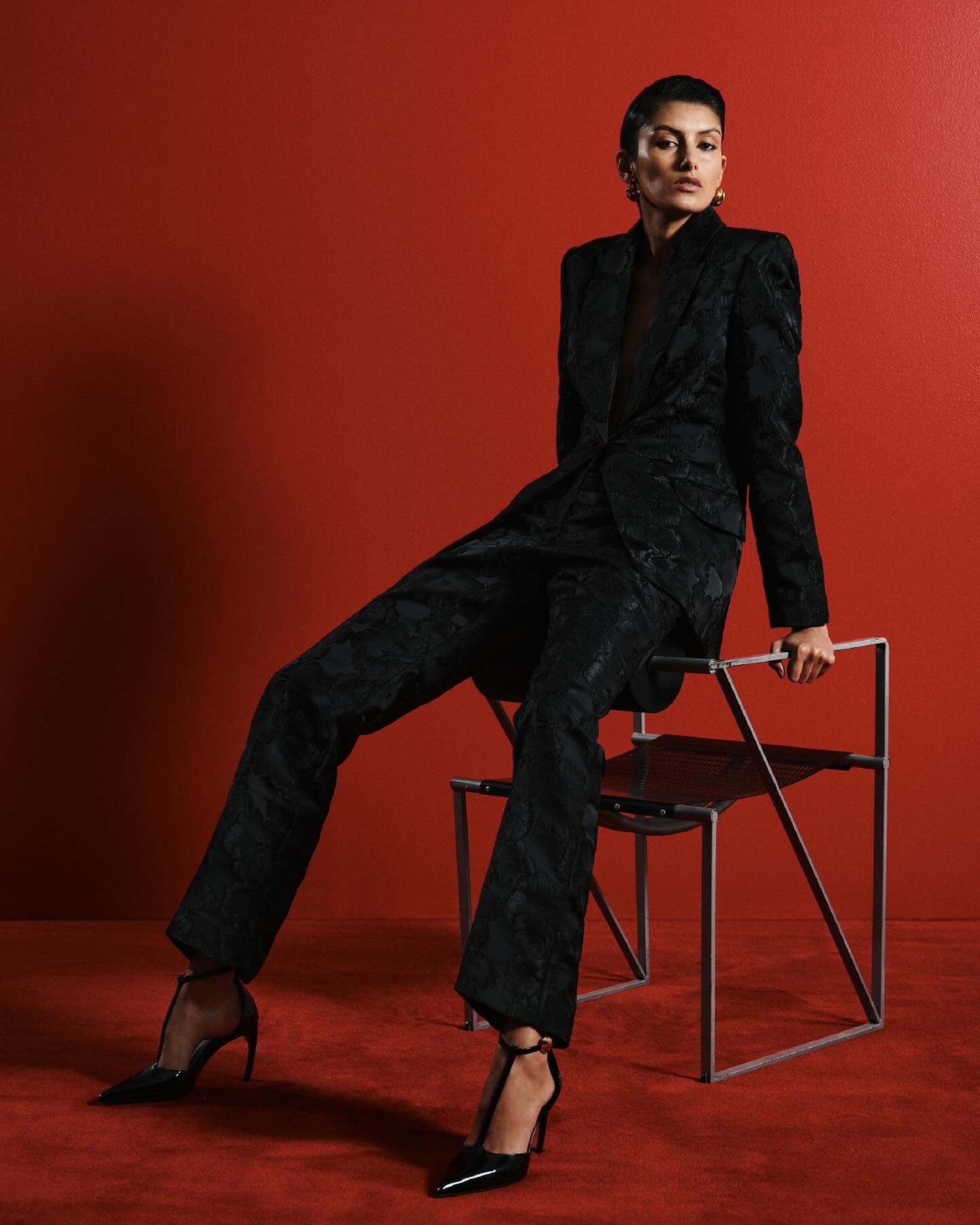 Woman in a black patterned suit poses while sitting on a modern chair against a red backdrop, wearing black high heels.