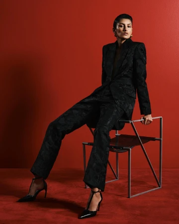 Woman in a black patterned suit poses while sitting on a modern chair against a red backdrop, wearing black high heels.