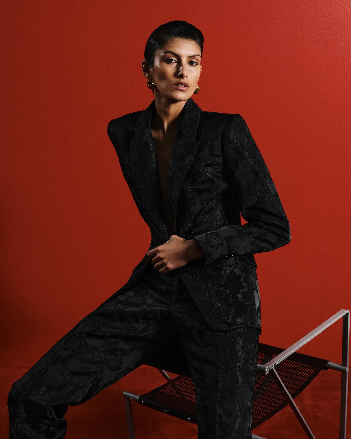 An adult female model poses in a black patterned blazer and pants, seated on a chair against a red backdrop in a fashion shot.