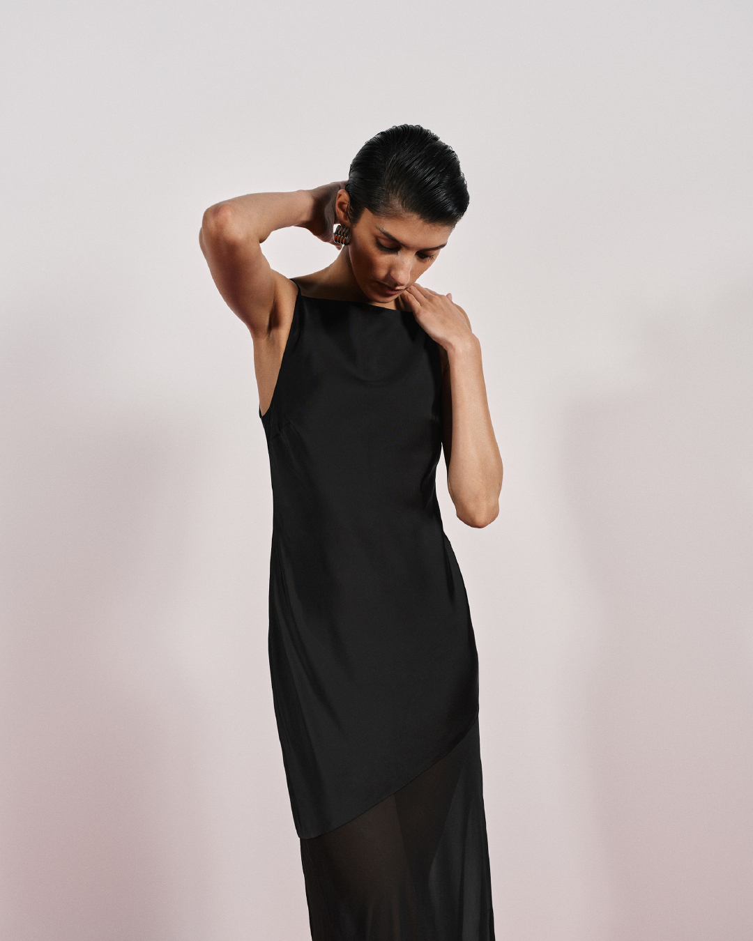 Model in a black slip dress with sheer hem posing against a plain light background for Cue Spring 24