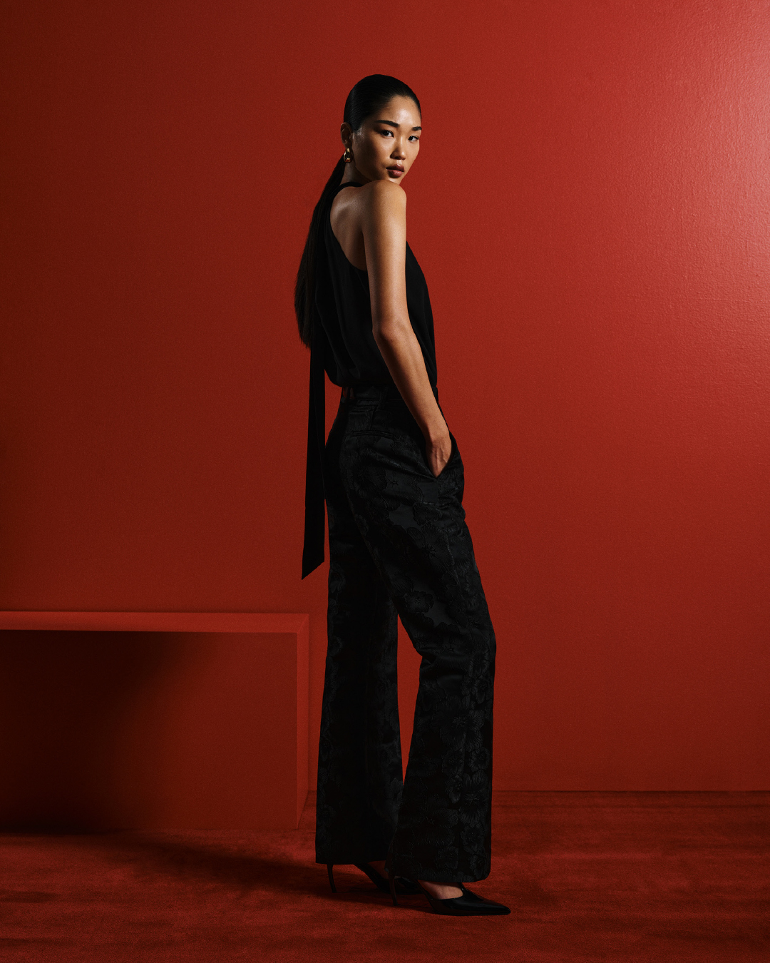 Model in black sleeveless top and patterned trousers standing against a vibrant red studio background for Cue Spring 24