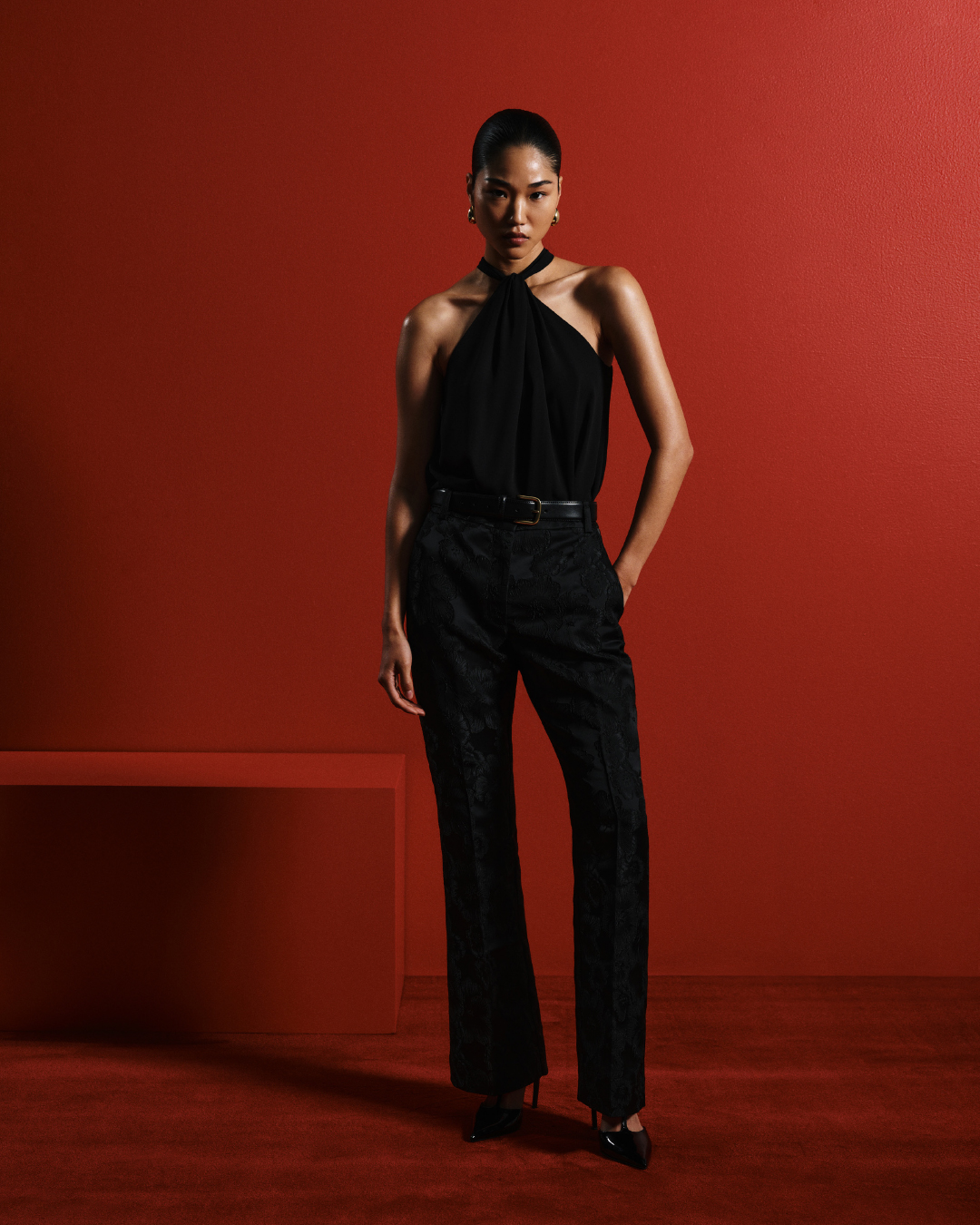 A woman in a black halter top and patterned black pants stands in front of a red wall, posing in a fashion shot with studio lighting.