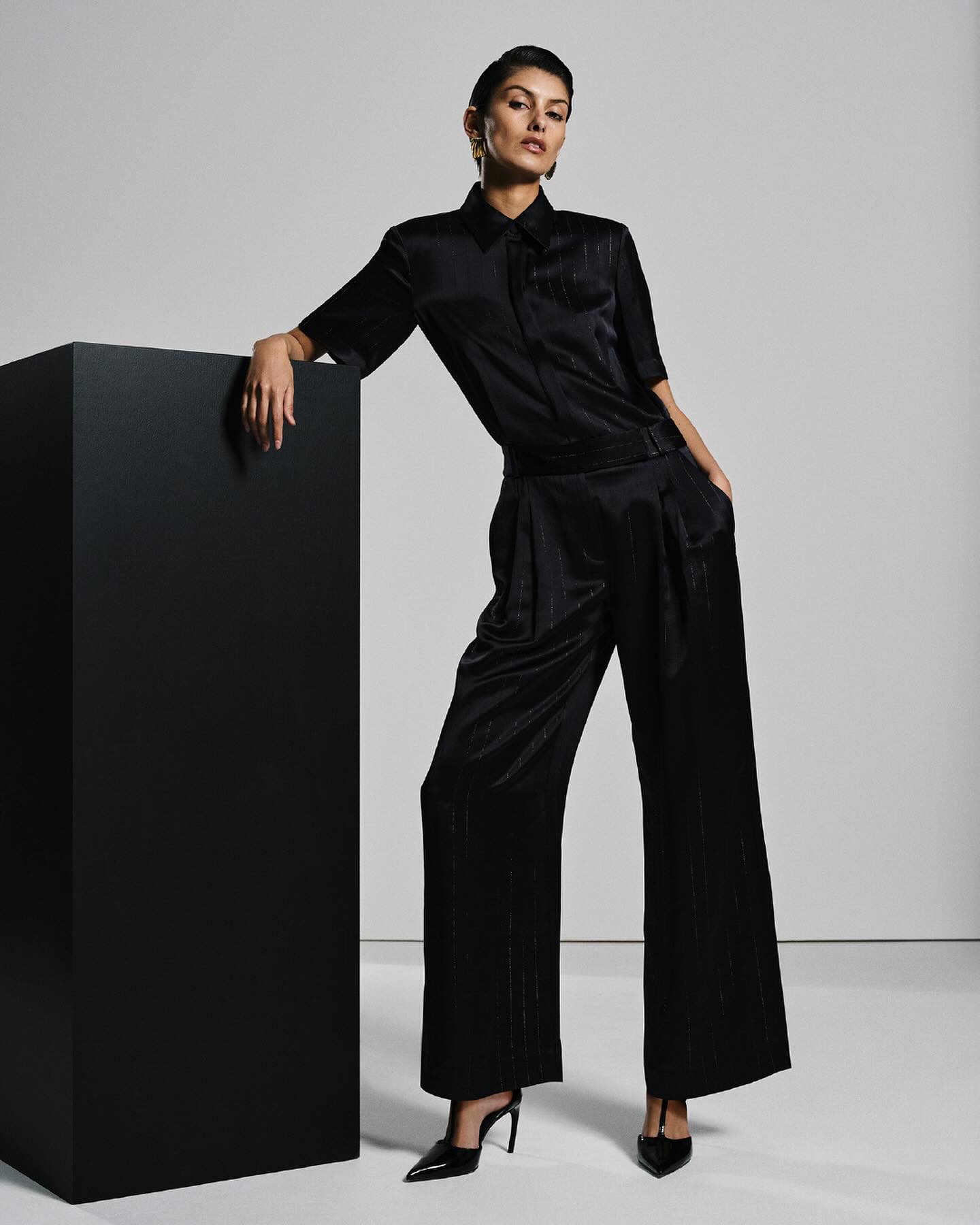 A woman in a dark silk shirt and wide-legged pants poses next to a black block, accented by pointed-toe heels and studio lighting.