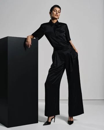 A woman in a dark silk shirt and wide-legged pants poses next to a black block, accented by pointed-toe heels and studio lighting.