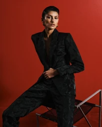 Model in a black patterned suit sitting on a chair against a bold red background for Cue Spring 24.