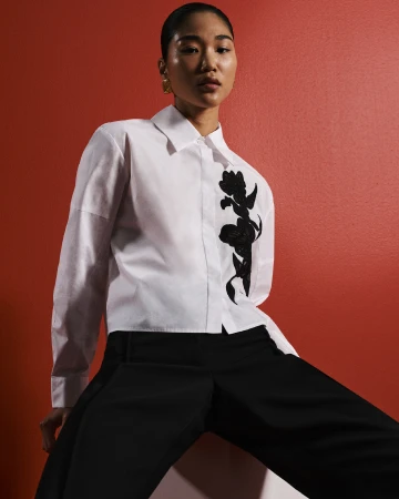 Model in white button-up shirt with black floral appliqué and black trousers against red backdrop for Cue Spring 24.
