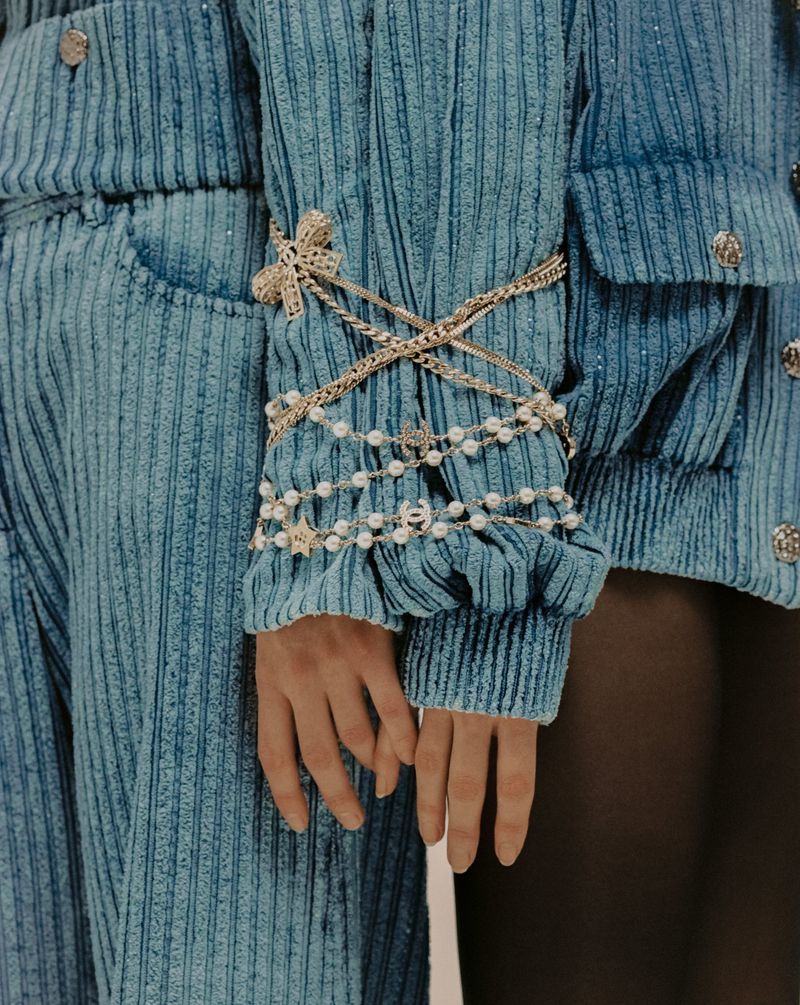 Close-up fashion shot of a woman's arm adorned with pearls and gold chains, wearing a textured blue jacket and pants.