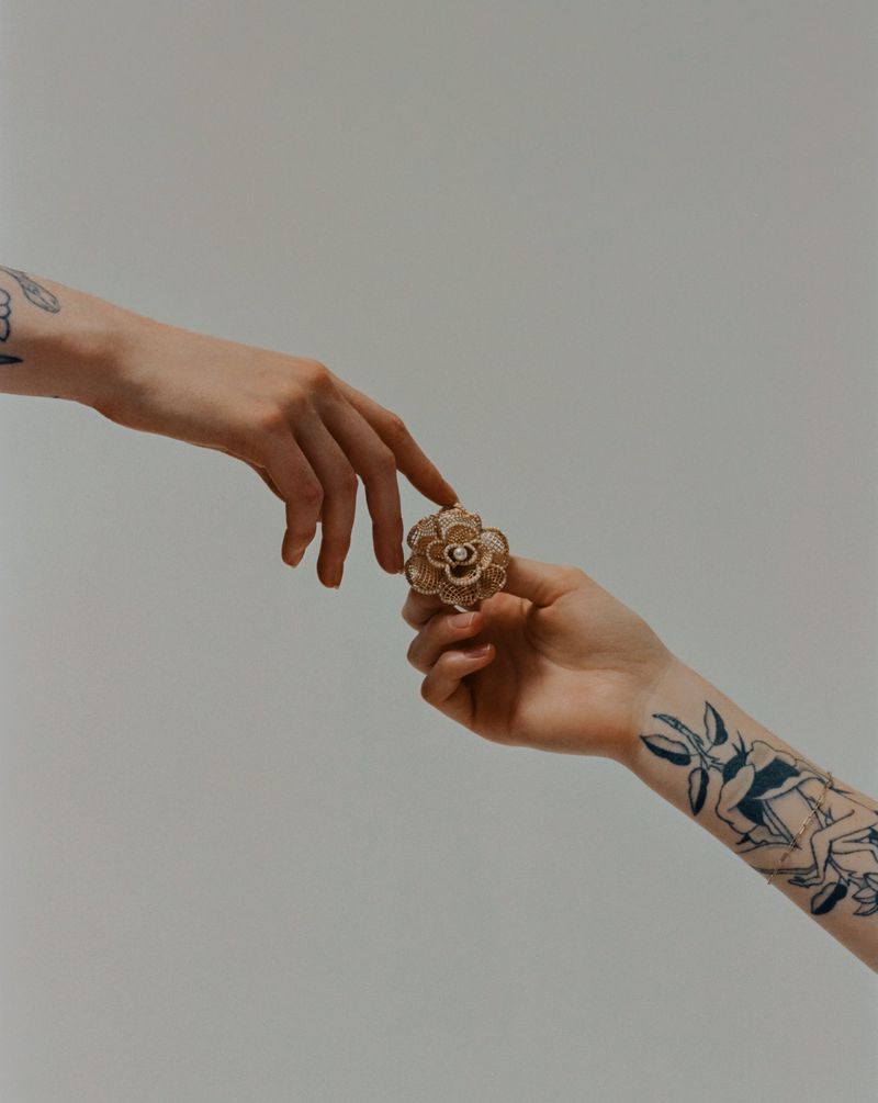 Close-up of a flower ring being passed between two hands with visible tattoos in a brightly lit indoor setting with a light background.