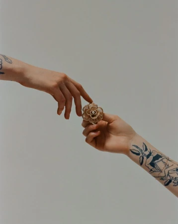 Close-up of a flower ring being passed between two hands with visible tattoos in a brightly lit indoor setting with a light background.