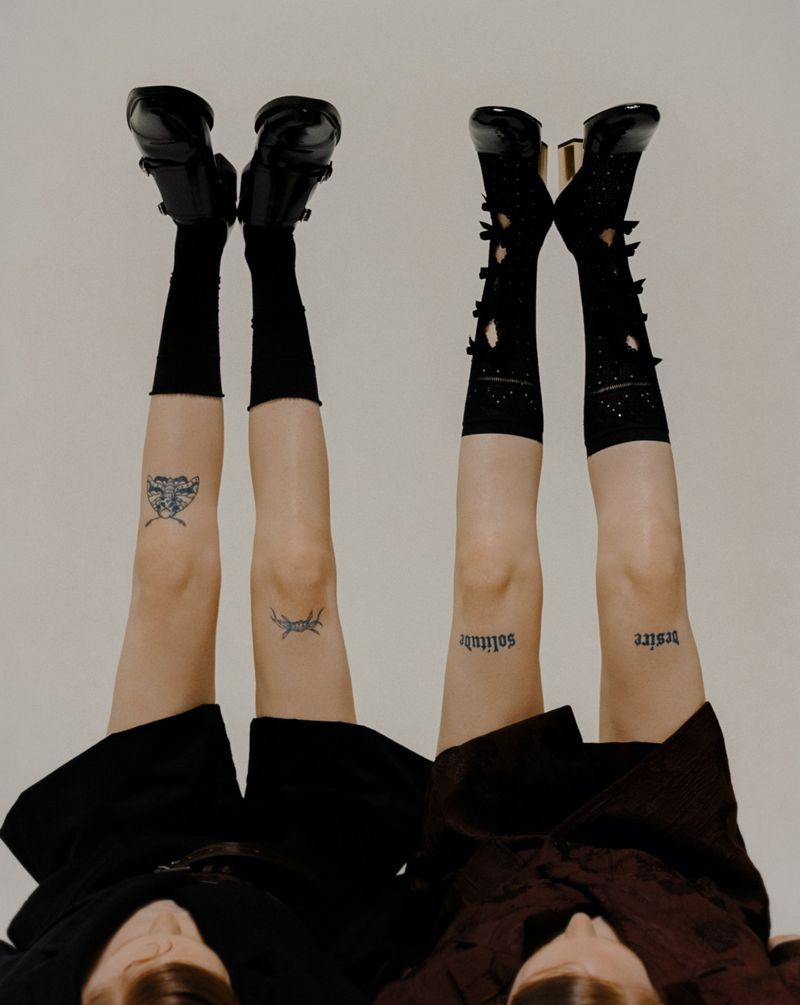 Two women lying upside down showcasing black Mary Jane shoes, knee-high socks with cutout details and tattoos on their legs.