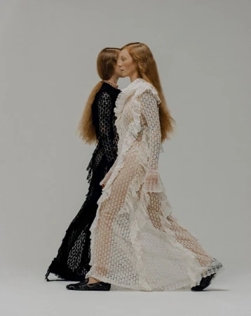 Two women wearing long textured dresses in a studio setting. One woman wears a white dress while the other wears a black dress.