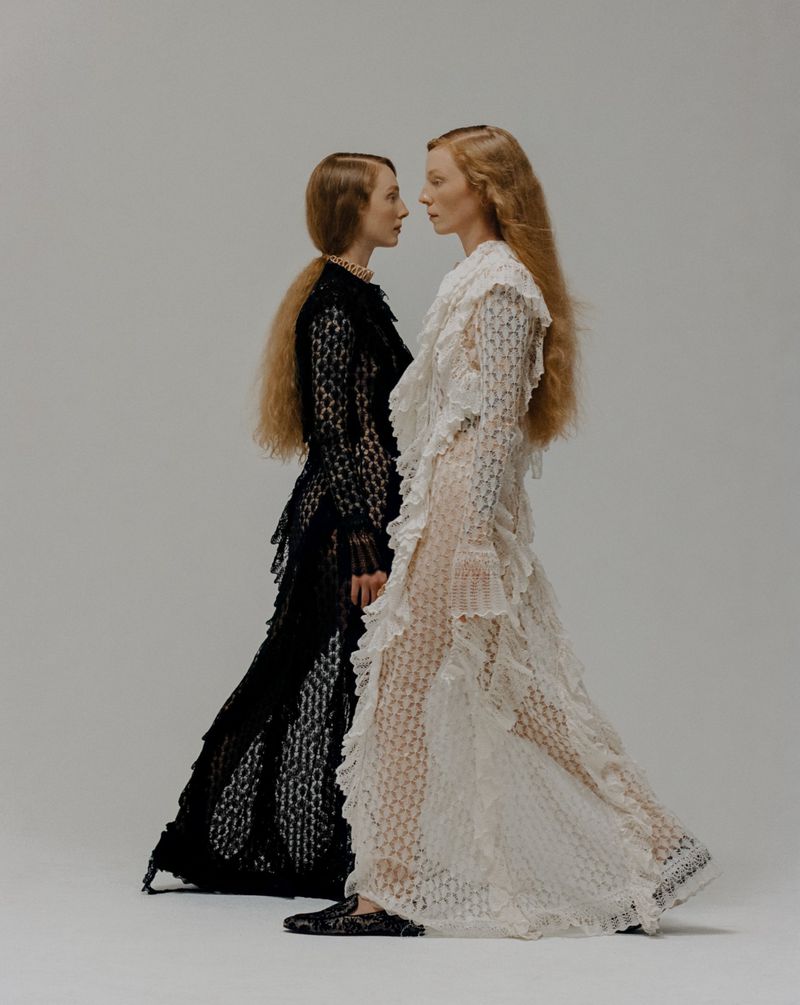Two women stand face-to-face in a studio wearing contrasting white and black gowns with textured frills and similar hairstyles.