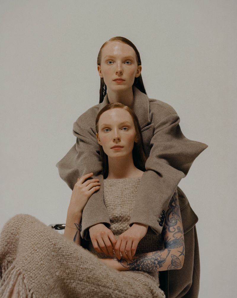 Two women in a photo studio. One is seated in front, the other stands behind with her arms around the seated woman's shoulders.