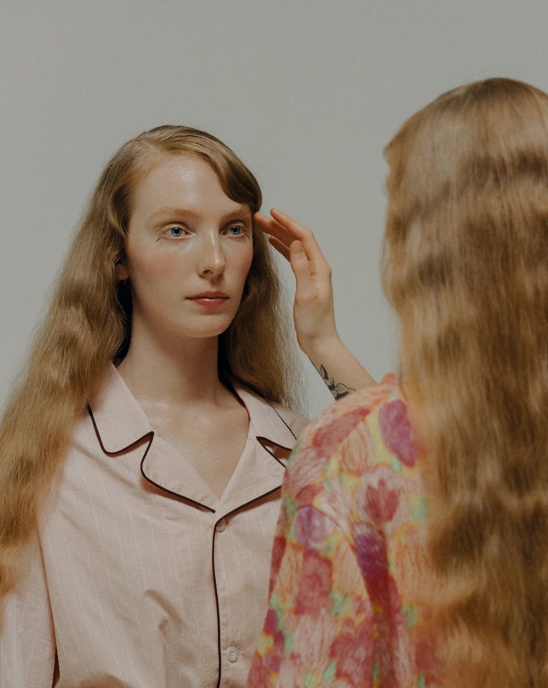 A woman with long red hair in pink pajamas touches another woman's hair in a light, neutral studio setting, portrait orientation.