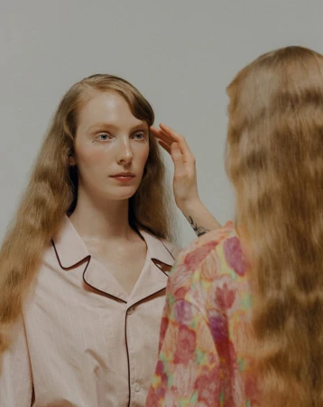 A woman with long red hair in pink pajamas touches another woman's hair in a light, neutral studio setting, portrait orientation.