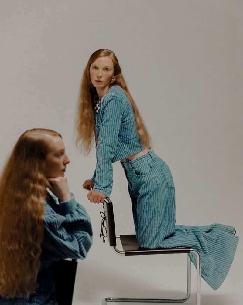 Two red-haired female models pose in matching blue striped outfits in a studio setting, one kneeling on a metal-framed chair.