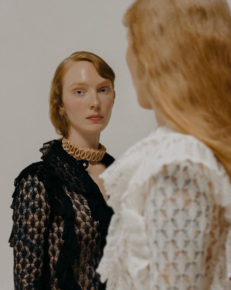 Two red-haired women in elegant lace dresses, one looking at the camera with a neutral expression against a plain backdrop.