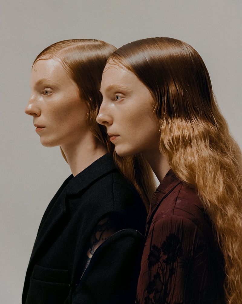 Two women stand side-by-side in a fashion portrait with slicked-back hair and minimalist style against a plain, light backdrop.