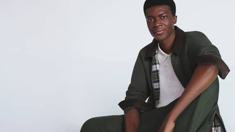 Portrait of a young Black man sitting and posing in an olive-green jacket with brown collar and cuffs, wearing a white ribbed shirt.