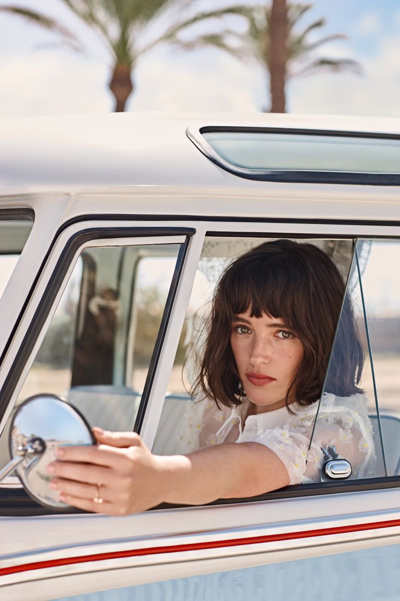 A brunette woman sits in a vintage car, gazing out the window while holding the side mirror in a sunny outdoor setting.