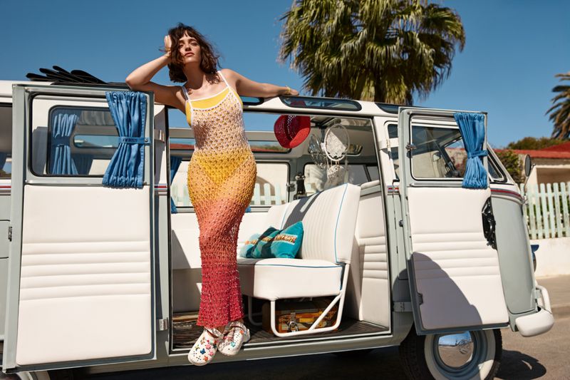 Woman in crochet dress leans against open door of a grey and white vintage van with a palm tree and blue sky in the background.