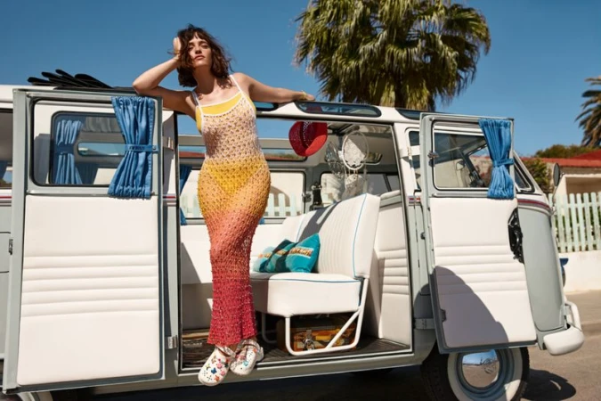 Woman in crochet dress leans against open door of a grey and white vintage van with a palm tree and blue sky in the background.