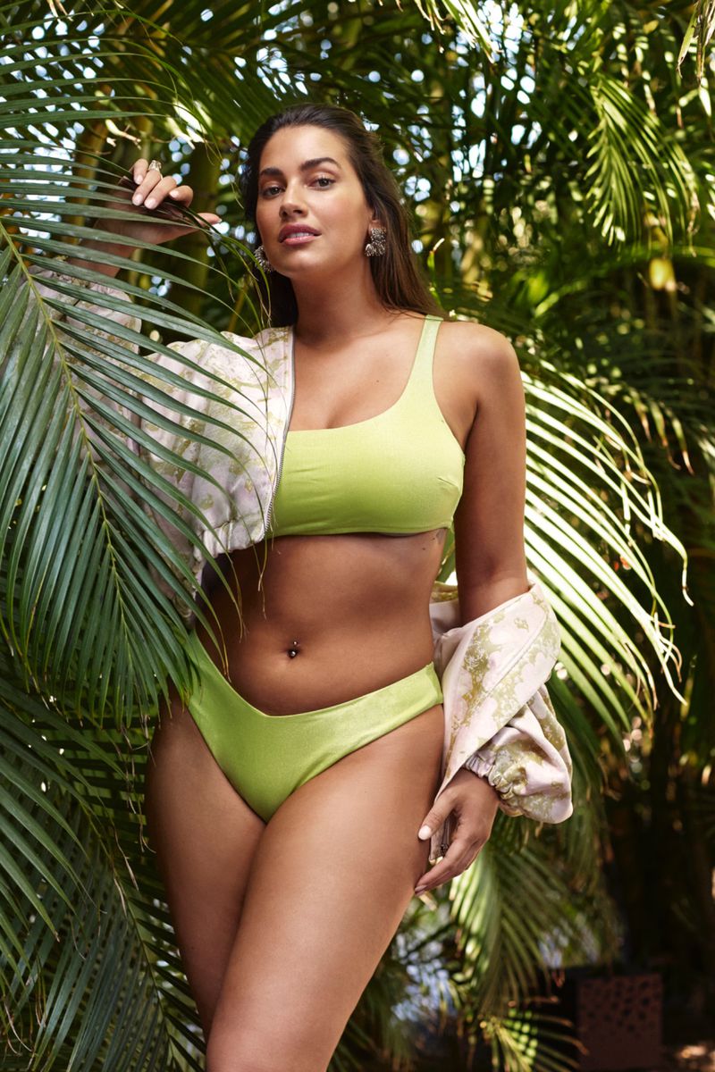Woman in lime green bikini poses in front of lush tropical foliage, fashionably draped with a textured jacket. Outdoor setting, natural light.