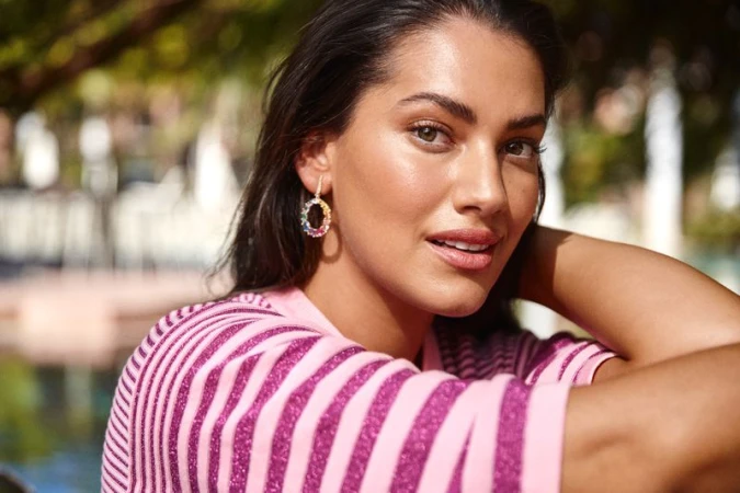 Portrait of a smiling woman with an oval multicolor gemstone earring, wearing a pink striped top in an outdoor setting with natural light.