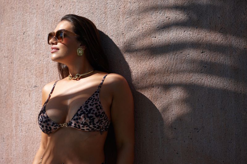 Adult female in bikini and sunglasses leaning against a wall in sunlight with shadows, accessorized with gold earrings and a necklace.