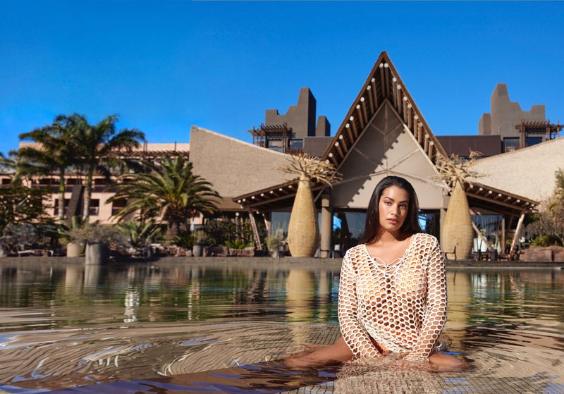Woman in a crochet dress sits in shallow water, with a resort building and palm trees visible under a clear blue sky.