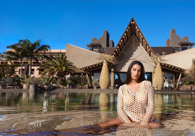 Woman in a crochet dress sits in shallow water, with a resort building and palm trees visible under a clear blue sky.
