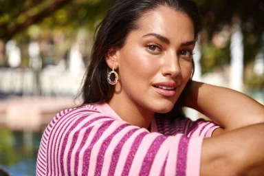 Model wearing a pink and purple striped top and multi-colored gemstone hoop earrings posing outdoors.