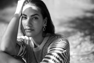 Black and white portrait of a woman wearing a striped top with her hand raised to her head outdoors.