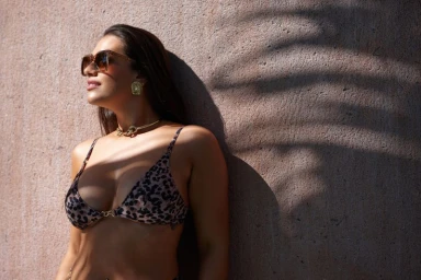 Model Lorena Duran wearing leopard print bikini and sunglasses leaning against textured stone wall